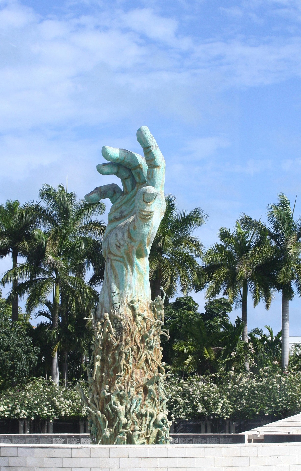 A large statue of a hand reaching up surrounded by smaller figures, in front of palm trees and a blue sky.